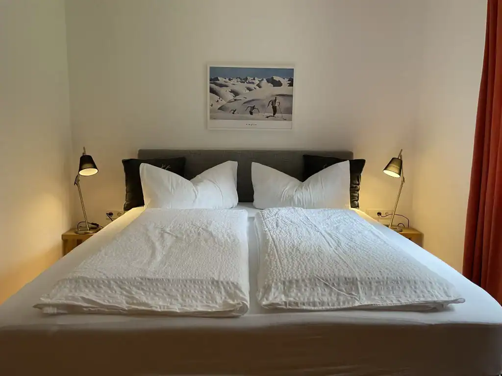 Doppelbett im Schlafzimmer mit weißen Bettwäsche, seitlichen Nachttischen und Wandbild mit Schneelan.